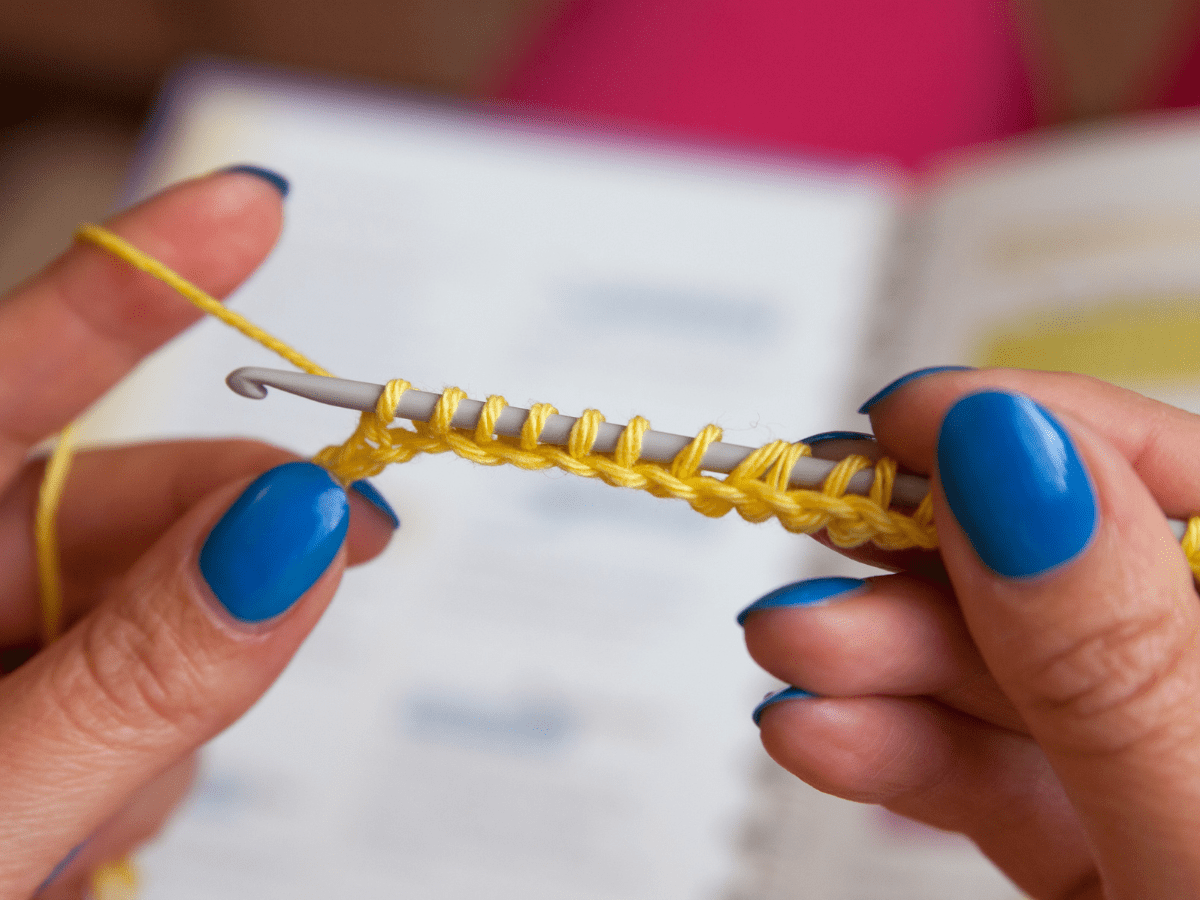 Hands with blue nail polish holding a crochet hook working yellow yarn in front of an open book